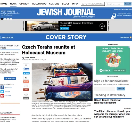 Several Czech Torahs, covered in ornate, colorful mantles and topped with decorative crowns, are lined up on a table inside a Holocaust Museum, featured as the main image in a Jewish Journal article about efforts to reunite these treasures.