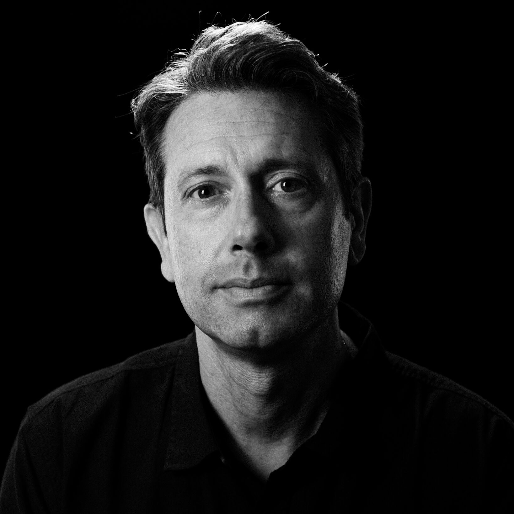 Black and white portrait of Nico S., a man with short hair, wearing a dark shirt, looking directly at the camera with a neutral expression against a dark background.