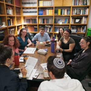 Eight people sit around a wooden table in a library or study room, engaged in discussion with papers, books, and drinks in front of them. Floor-to-ceiling bookshelves filled with books line the walls.