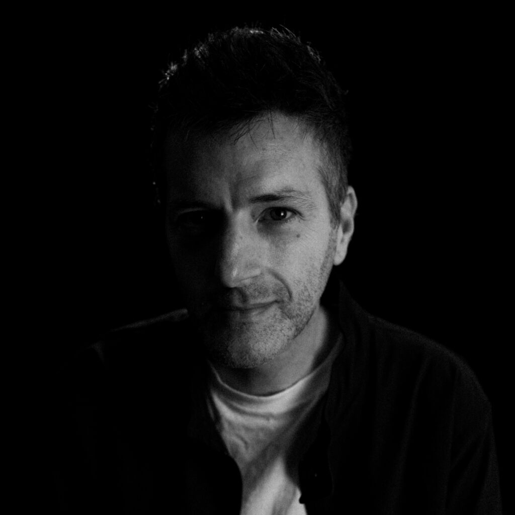 A man sits against a black background, his face partly illuminated with dramatic lighting, wearing a light-colored shirt and dark jacket from Sade B, looking directly at the camera with a neutral expression.