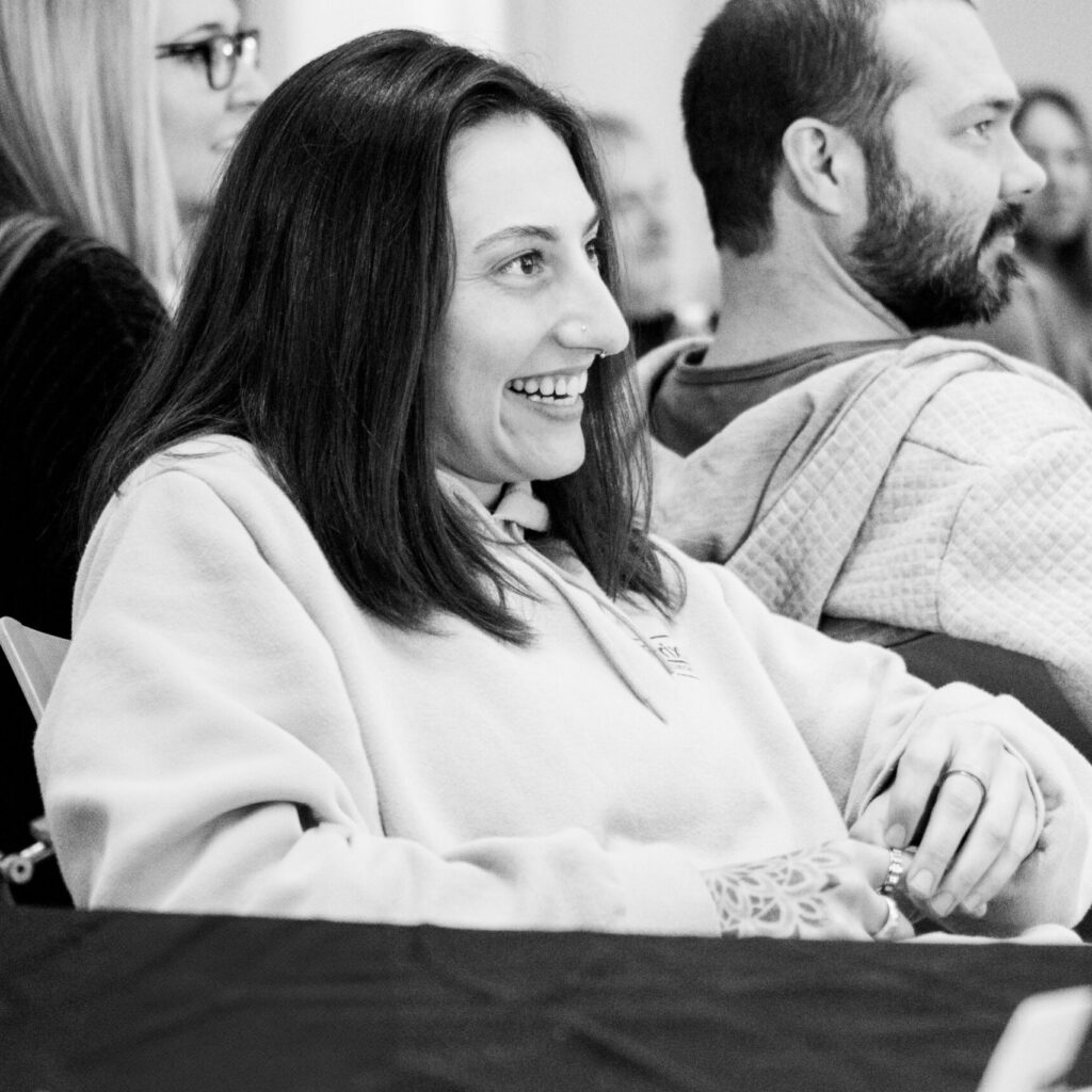 A woman smiling and sitting at a table with others in a casual indoor setting, possibly discussing rehab or how to find a treatment center you can trust. The image is black and white, creating a relaxed and friendly atmosphere.