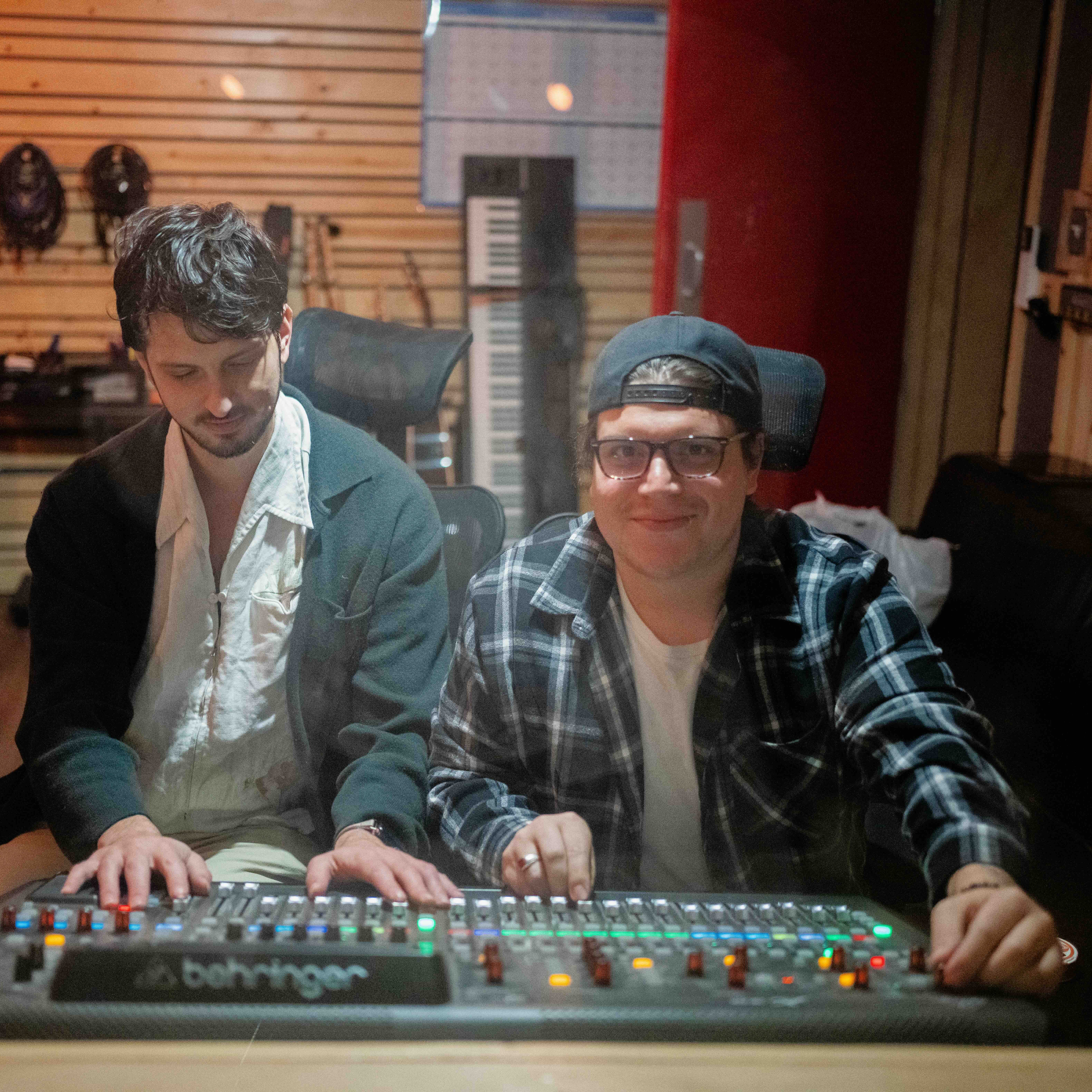 _DSC1600-2 Two men sit at a sound mixing board in a recording studio. One is adjusting the controls for their music program, while the other watches and smiles. The studio has wood paneling, audio equipment, and coiled cables on the wall.
