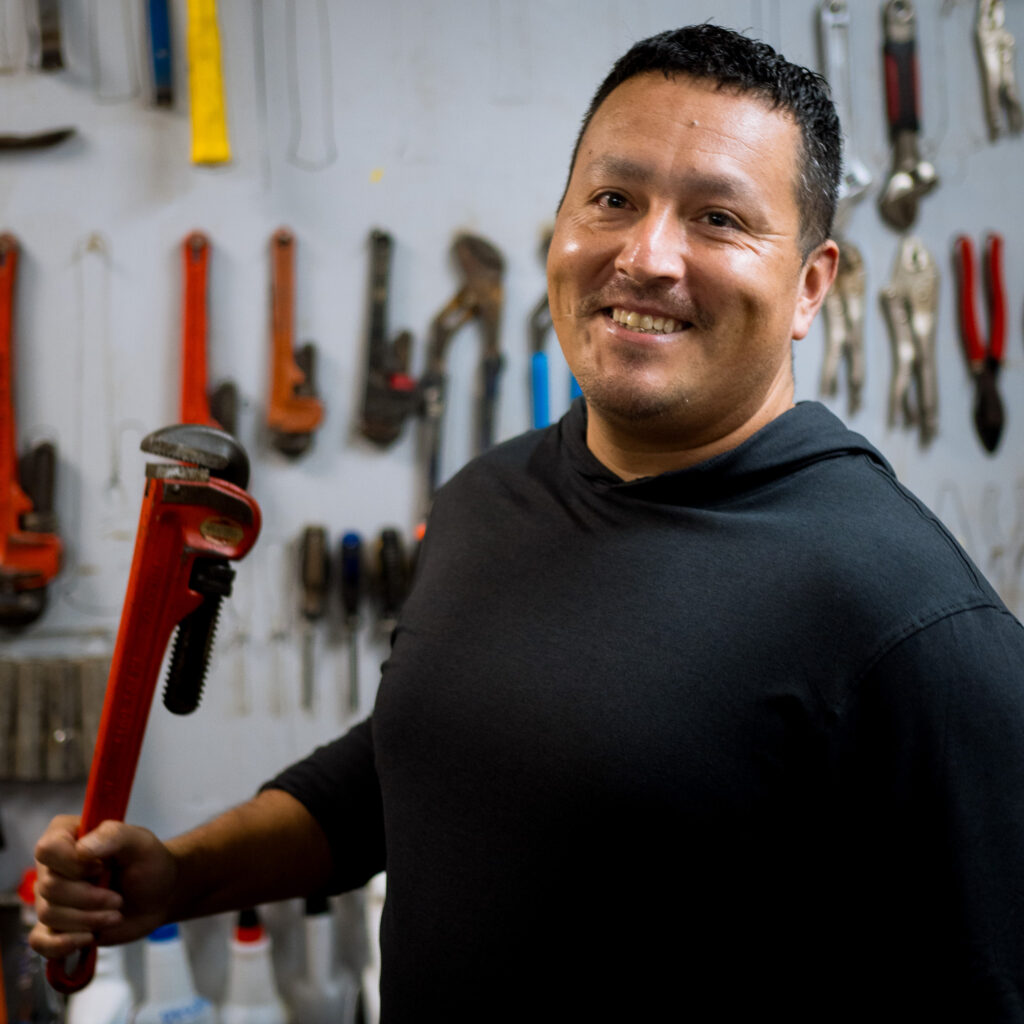 A smiling man in a black shirt holds a large red pipe wrench in a workshop, with various tools and a Klea p sign hanging on the wall behind him.