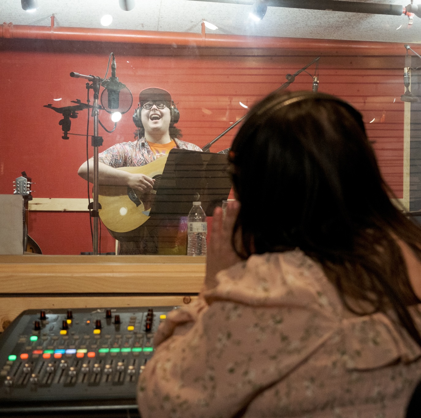 _DSF3130 A person sings and plays guitar in a recording studio, standing at a microphone as part of a Music Program. Another person, seen from behind, sits at a sound mixing board, watching through the studio glass.