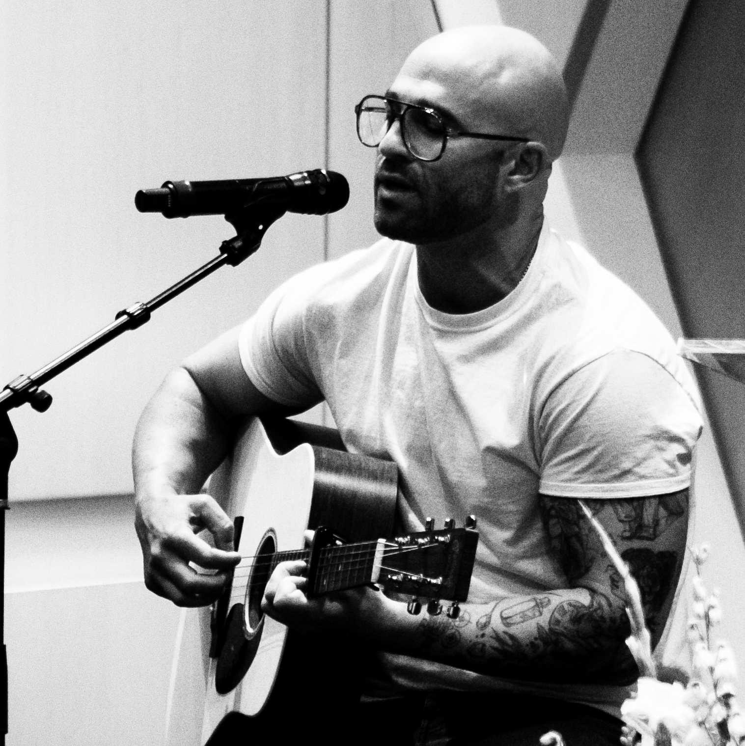 High Holidays ’25-111 A man with a shaved head, wearing glasses and a white t-shirt, plays an acoustic guitar and sings into a microphone as part of a music program. The black and white image captures his passion for performance.