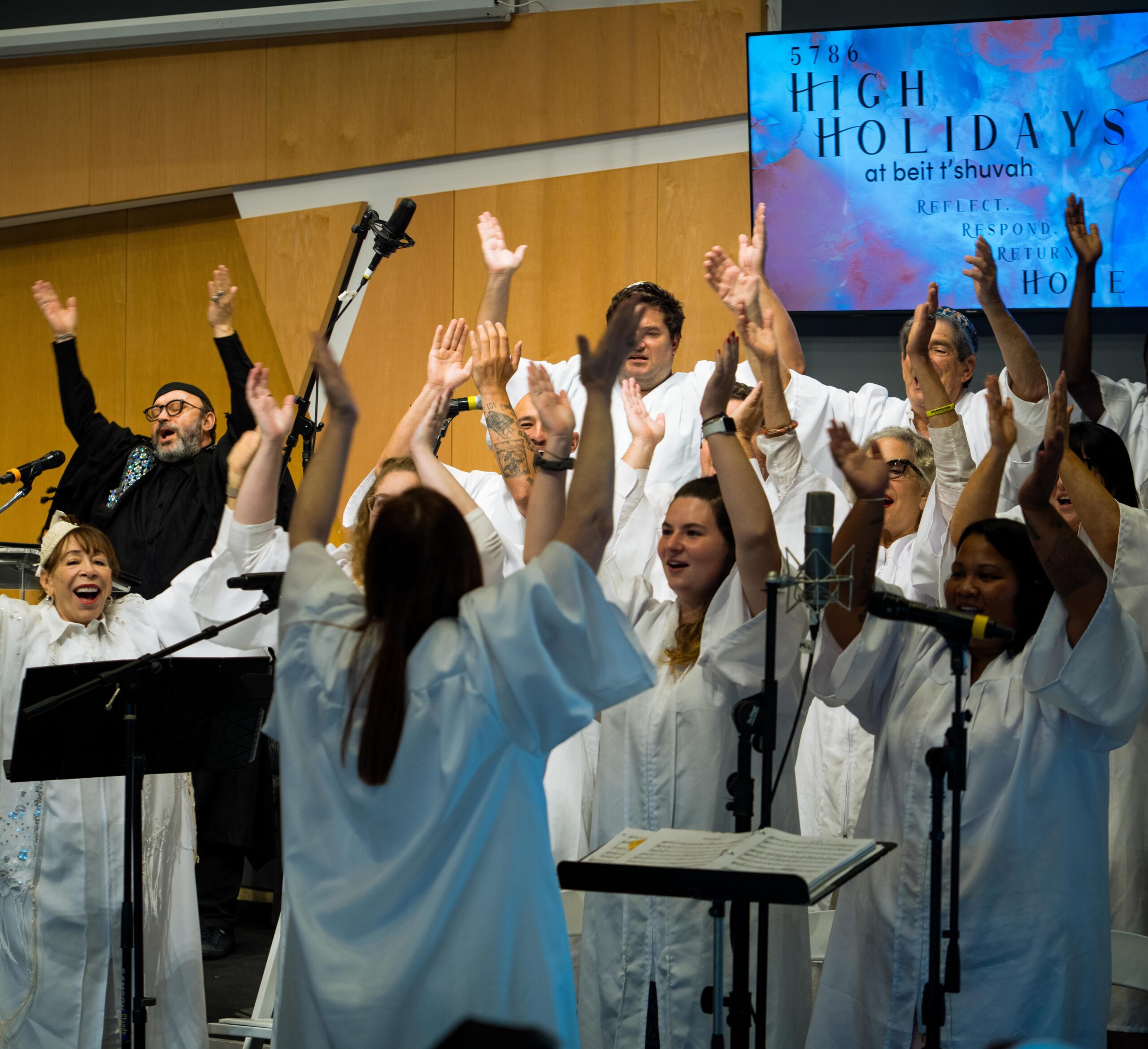 High Holidays ’25-135 A choir dressed in white robes sings joyfully with raised arms during a High Holidays service at Beit T’Shuvah, as seen on a sign in the background. The group is led by a conductor, highlighting the vibrant Music Program.