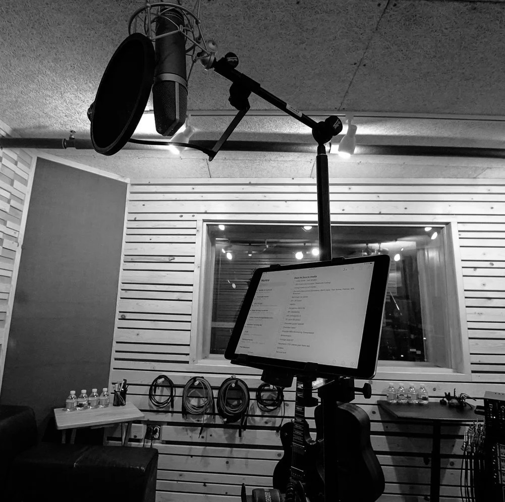 Studio+Photos.jpg copy A recording studio in black and white showcases a microphone, pop filter, and lyrics on a tablet stand—perfect for any music program. The background features a window, soundproof walls, coiled cables, and water bottles on a table.