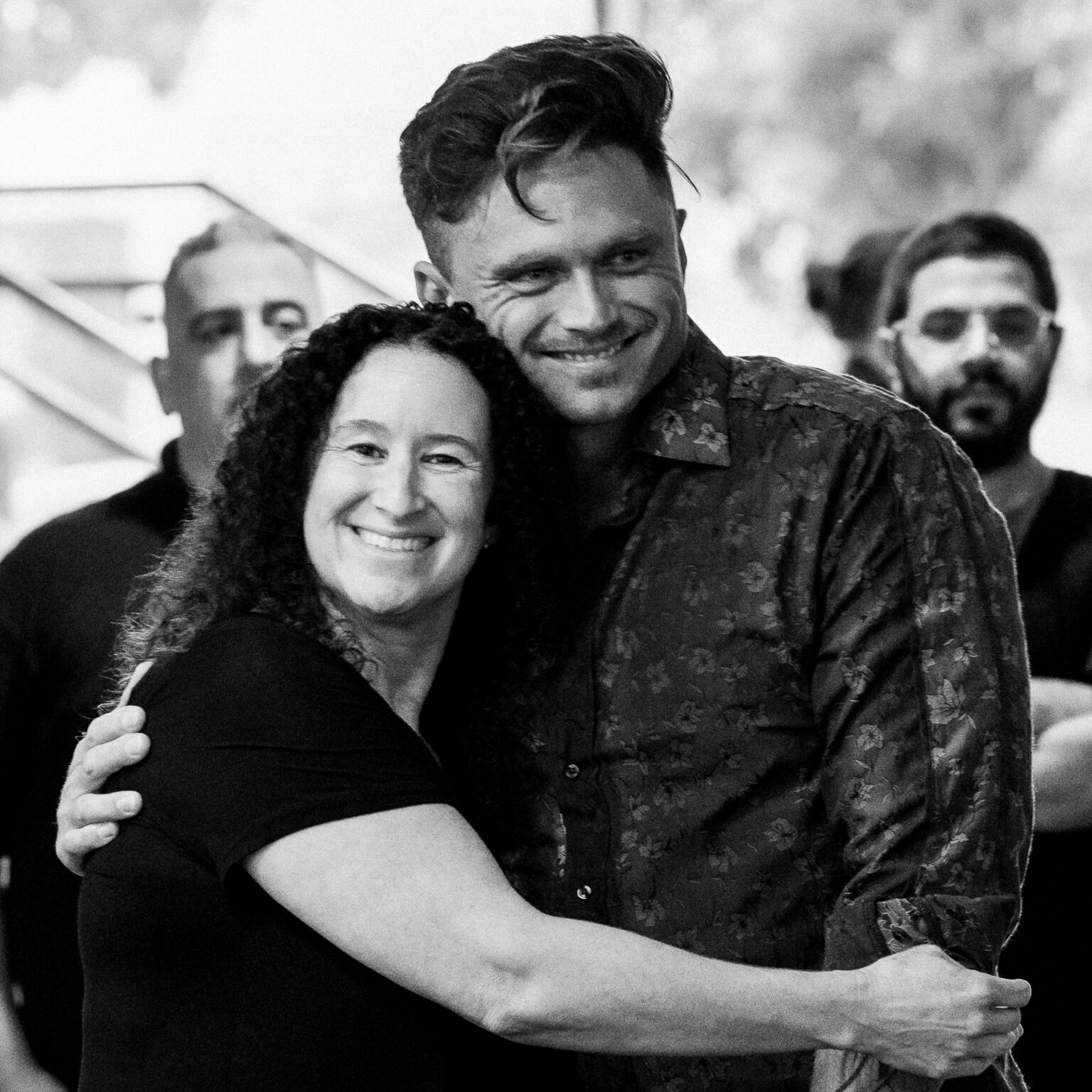 A woman and a man stand close together, smiling and hugging, celebrating their addiction recovery. They are surrounded by other people in the background, and the photo is in black and white.