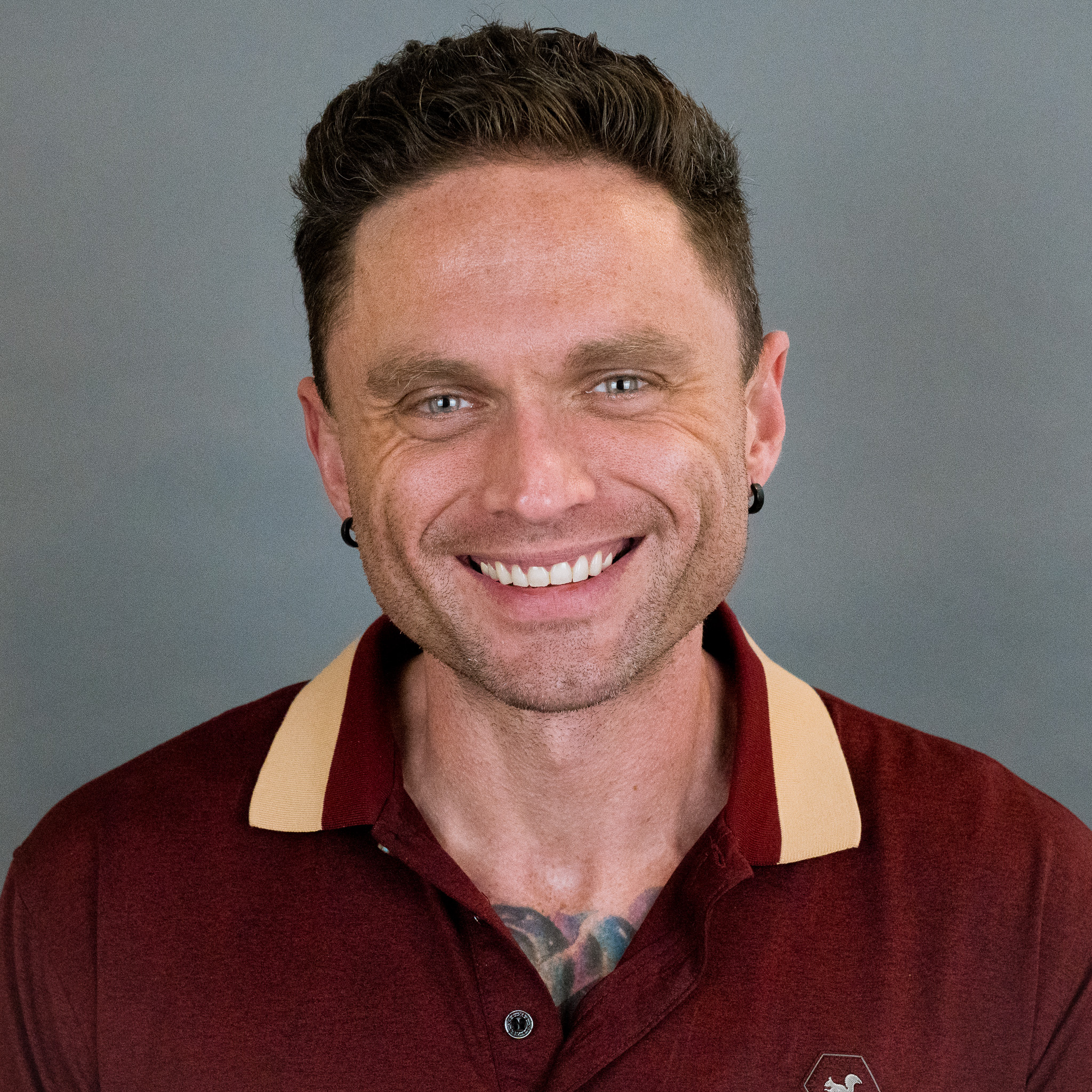 A smiling man with short curly brown hair, wearing a maroon collared shirt with tan accents and earrings, stands in front of a plain gray background, ready to collaborate with the Development Team.