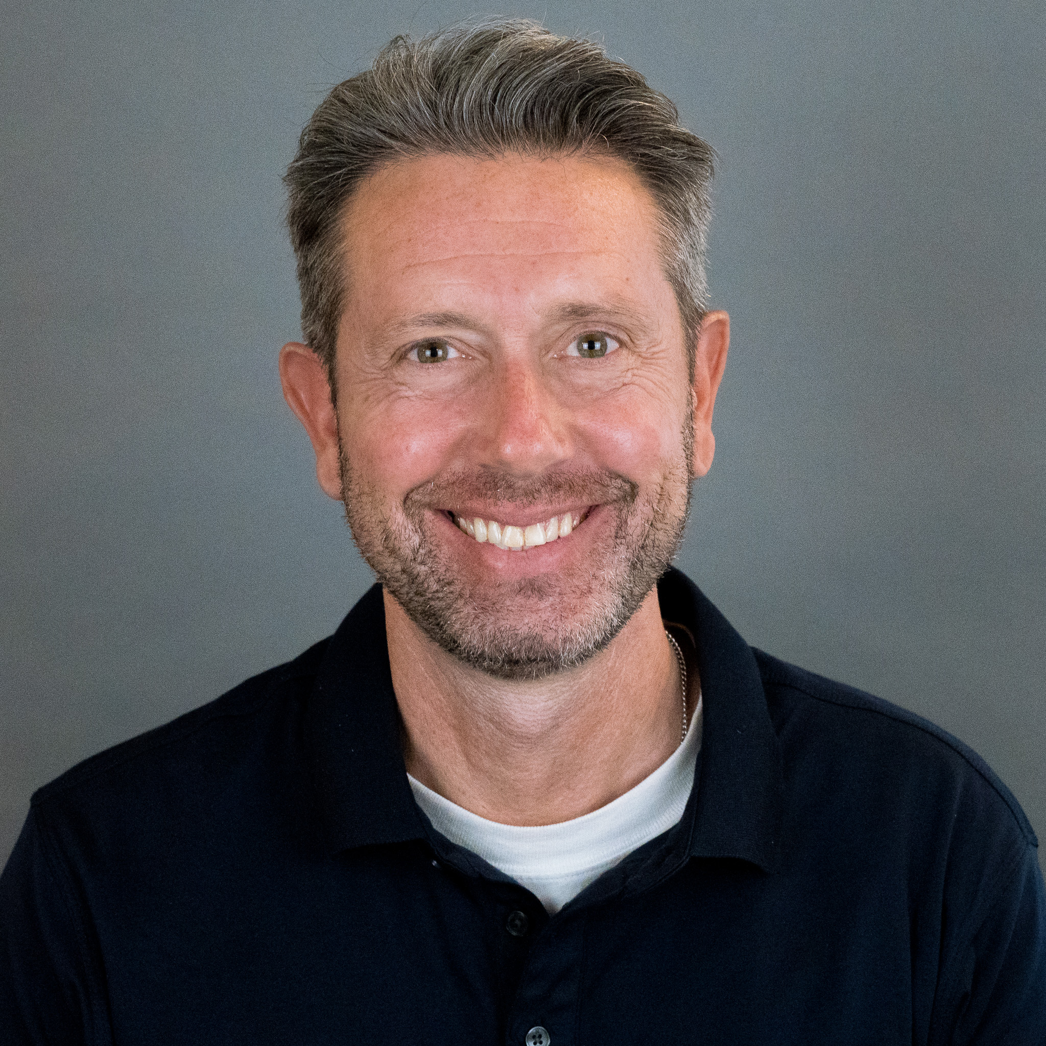 A middle-aged man with short gray hair and a beard smiles at the camera. He is wearing a black collared shirt over a white t-shirt, representing the Development Team against a neutral gray background.