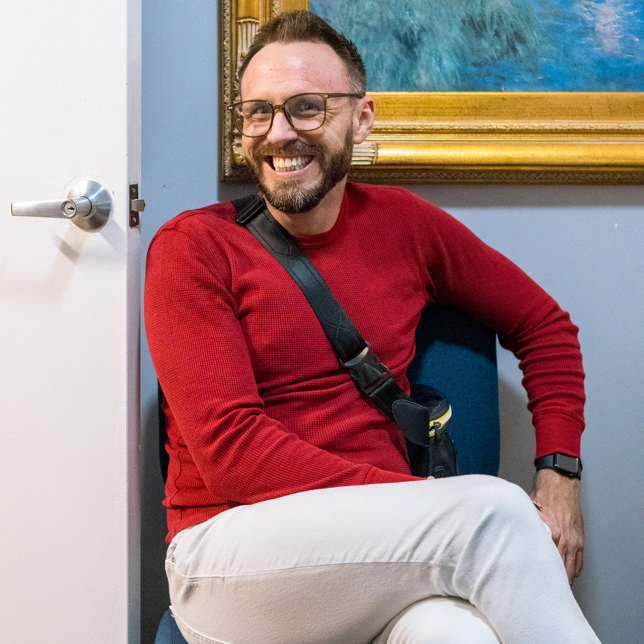 A man with glasses, a red sweater, and light pants sits on a chair with legs crossed, smiling. He has a crossbody bag and wears a watch, appearing relaxed—like he’s just finished an IOP or outpatient rehab session. A framed painting hangs behind him.