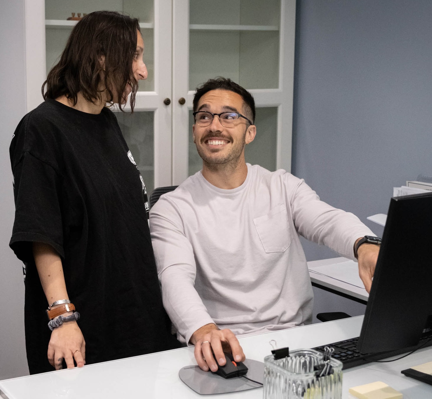A man sitting at a desk using a computer smiles up at a woman standing next to him, discussing their progress in the iop. They appear to be having a friendly conversation in an office setting.