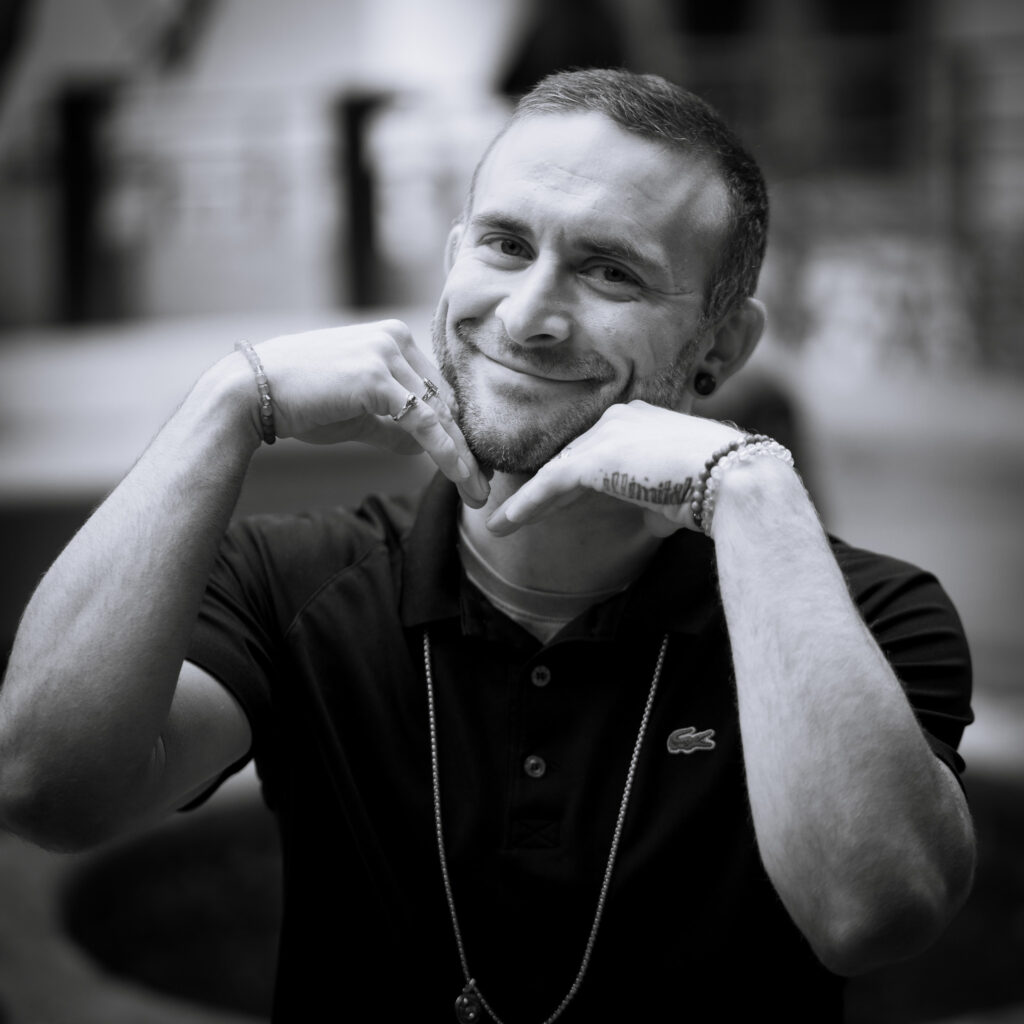 A man in a black polo shirt smiles playfully at the camera, resting his chin on his fists. The background is blurred, and the photo is in black and white, capturing a candid moment reminiscent of Rachel S’s signature style.