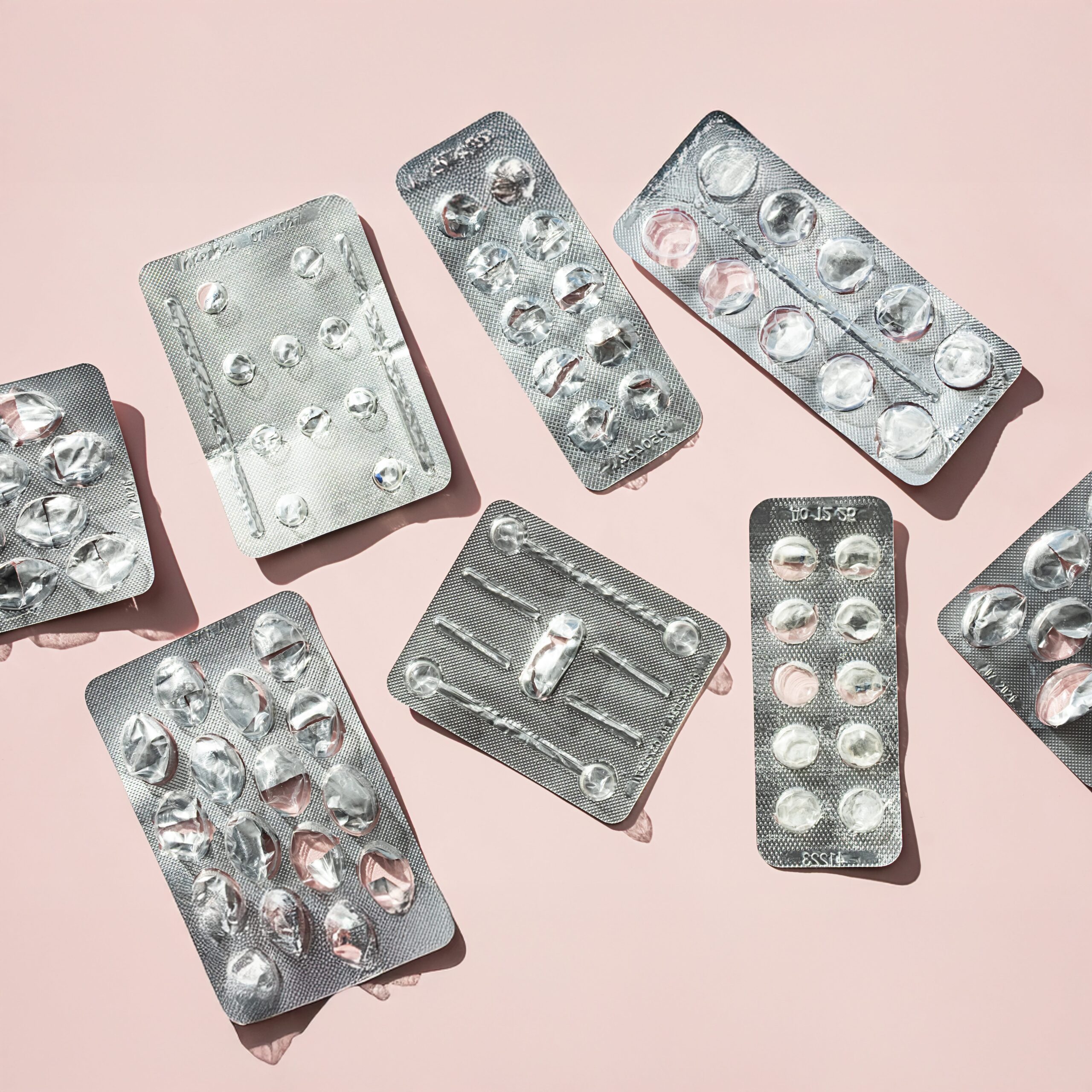 Several empty and partially empty blister packs of pills are scattered on a light pink surface, with most compartments showing missing tablets, highlighting Harm Reduction In Action: Medication Assisted Treatment Explained.