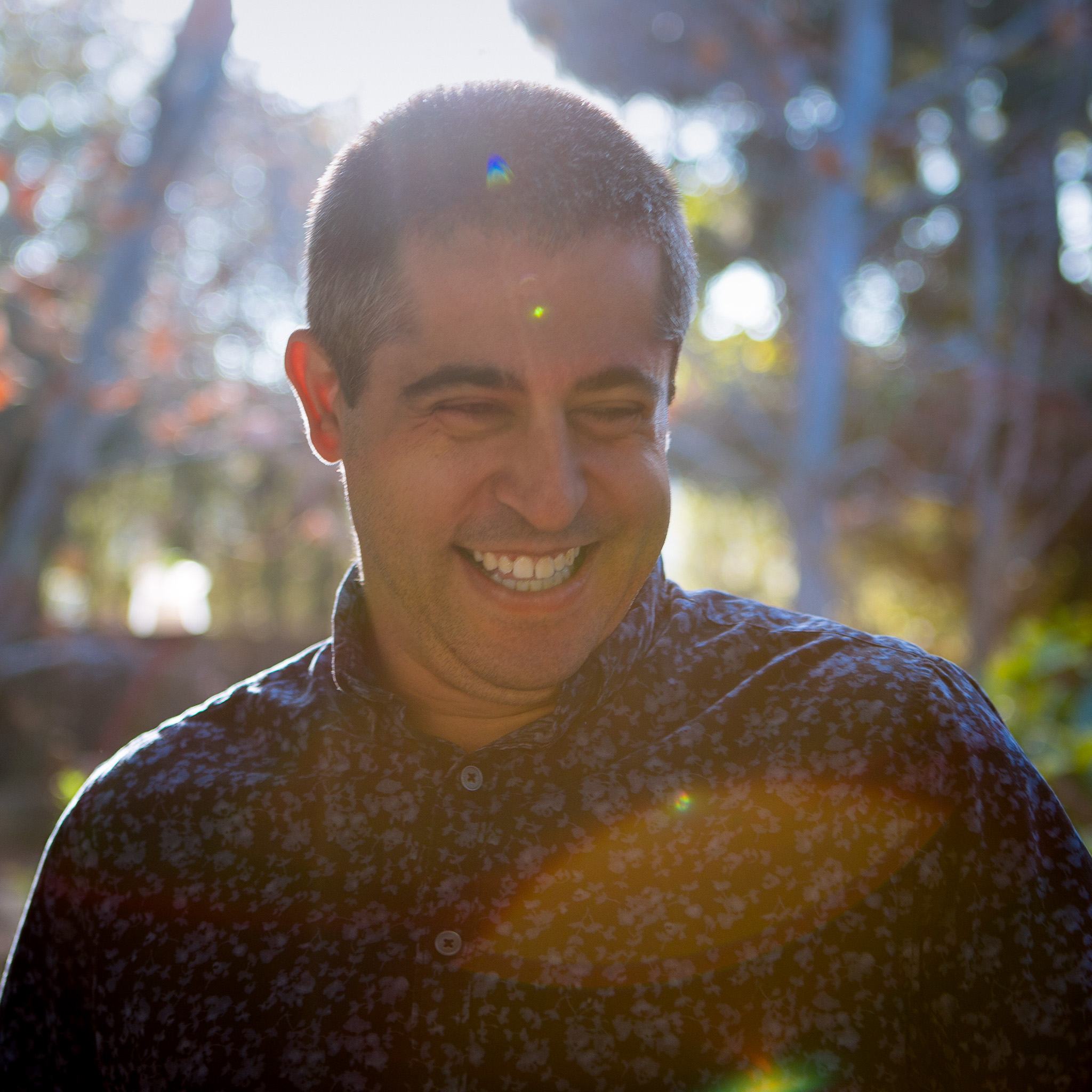 A man with short dark hair smiles brightly outdoors, sunlight and lens flare illuminating his face. He wears a dark, patterned shirt and stands in front of a blurred, natural background with trees—captured by Stacey G.