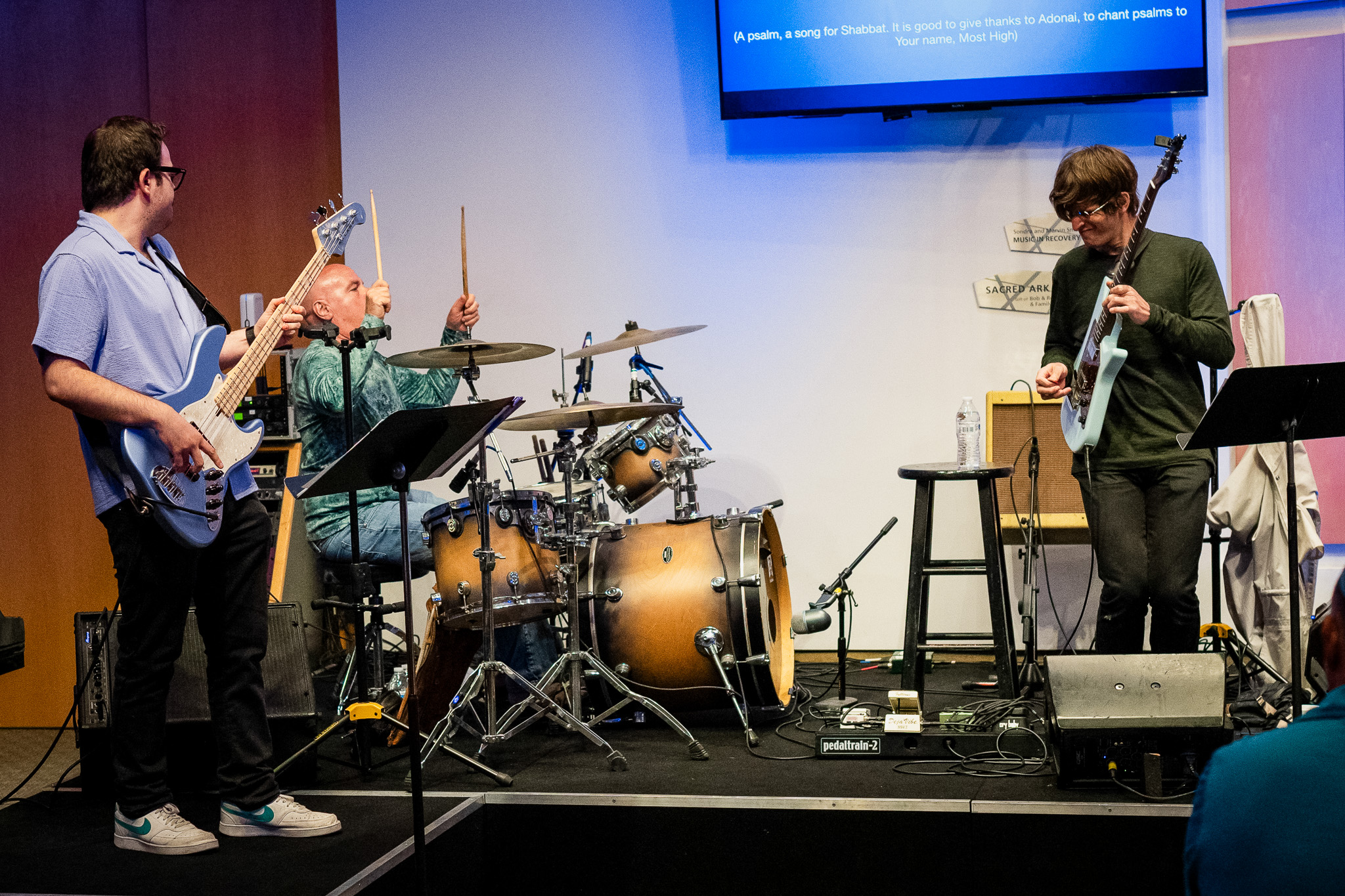 Three musicians perform on a small stage for a Temple Membership event: one on bass guitar, one on drums, and one on electric guitar. Sheet music stands are in front of them, with a screen displaying text above the stage.