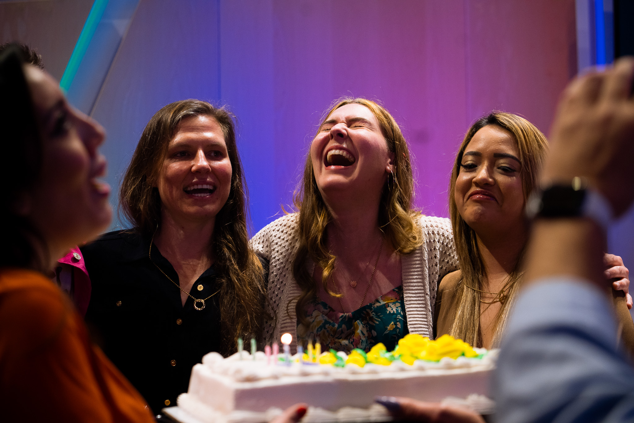Three women stand together, smiling and laughing, behind a birthday cake with lit candles. The festive scene reflects the joy and community often found when you join temple events and experience temple membership benefits.