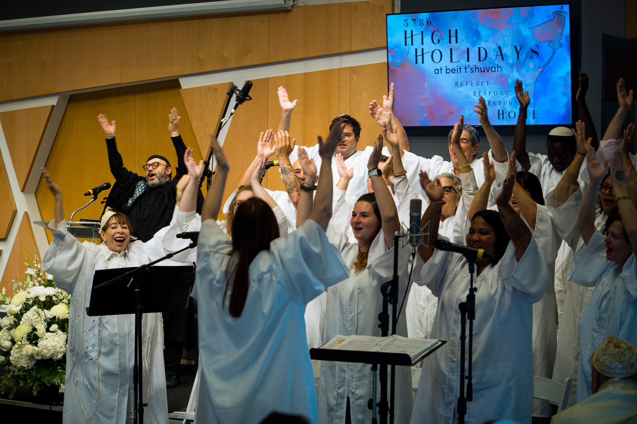 A diverse group of people in white robes joyfully sing and raise their arms on stage, celebrating the spirit of Temple Memberships. Behind them, a sign reads “HIGH HOLIDAYS at beit t’shuvah.” Microphones and music stands are in front of the choir.
