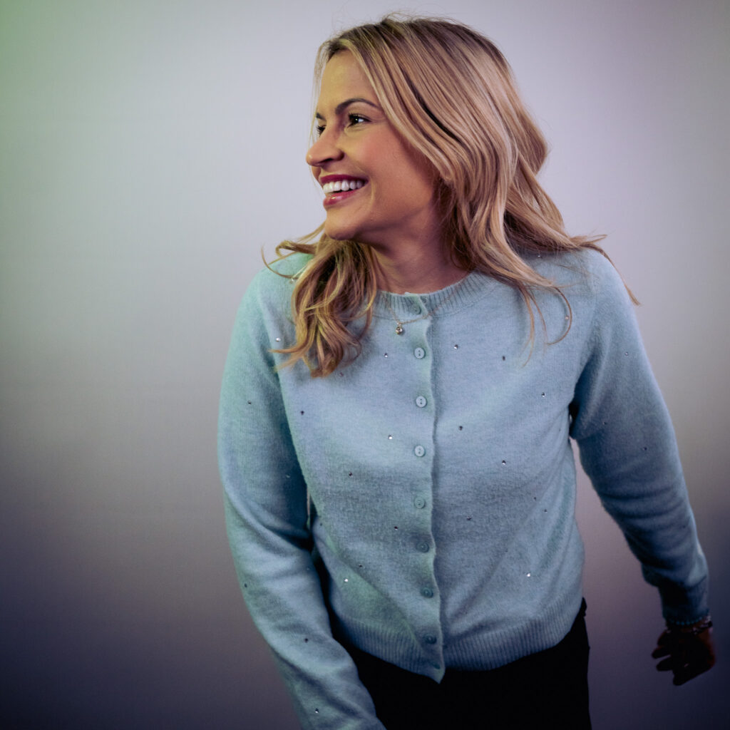 A woman with long blonde hair wearing a light blue cardigan smiles and looks to her left against a plain, softly lit background, in a portrait inspired by Agustin L.