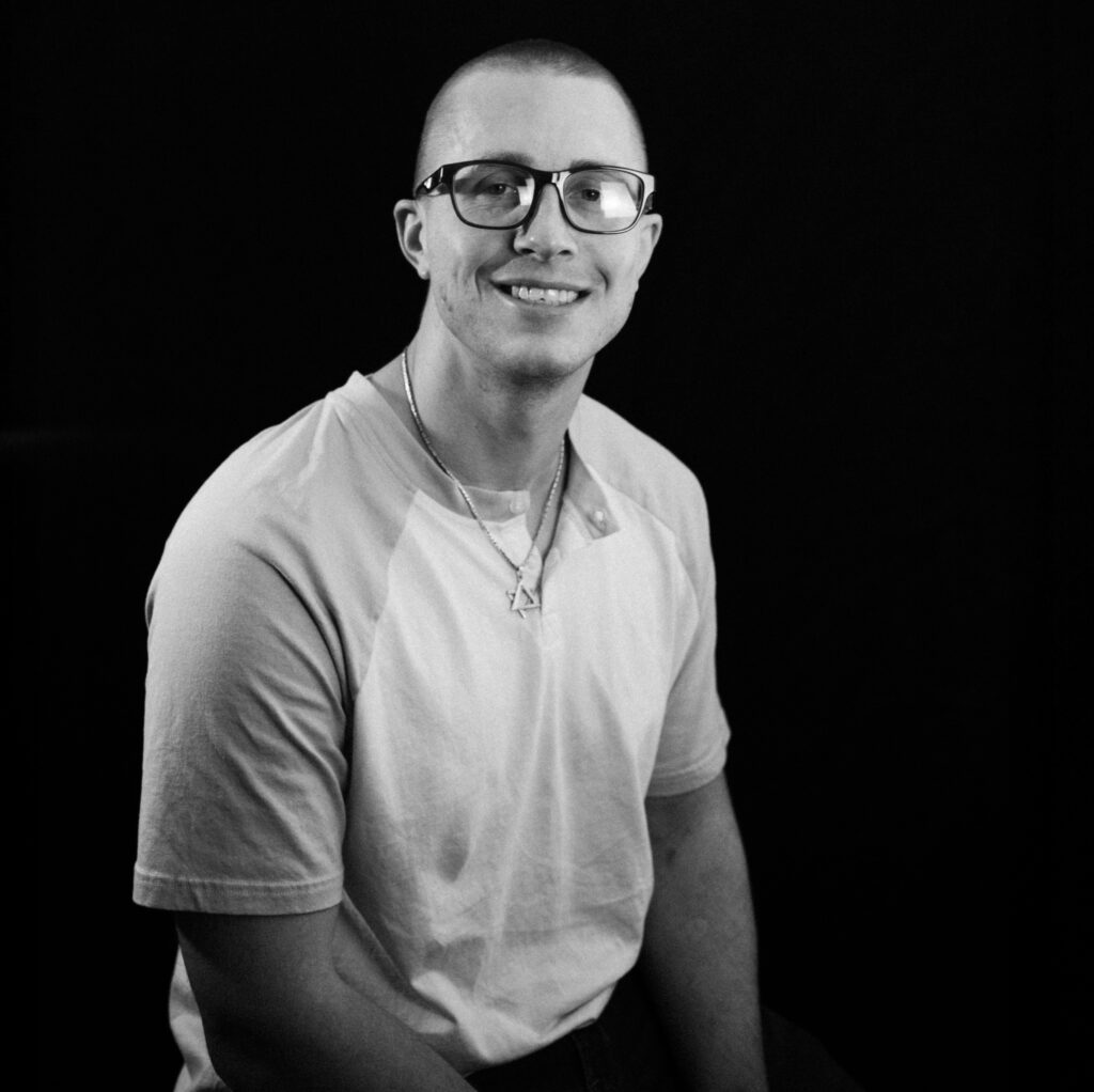 A young man with short hair and glasses, possibly Kyle P, smiles while sitting against a dark background. He is wearing a light-colored t-shirt and a necklace. The black and white photo highlights his relaxed, friendly expression.