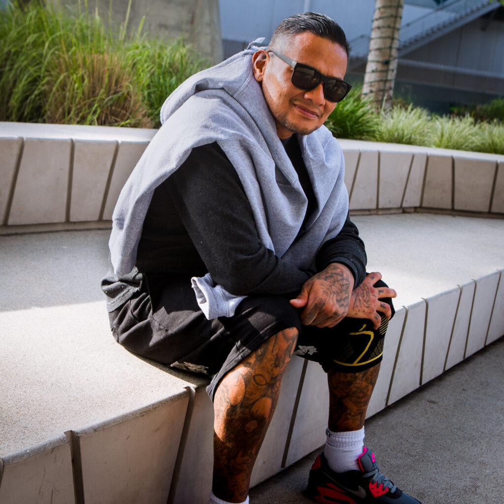 A man with tattoos, sunglasses, and a hoodie draped over his shoulders sits on a white outdoor bench, smiling at the camera on a sunny day—captured perfectly by Julia t.