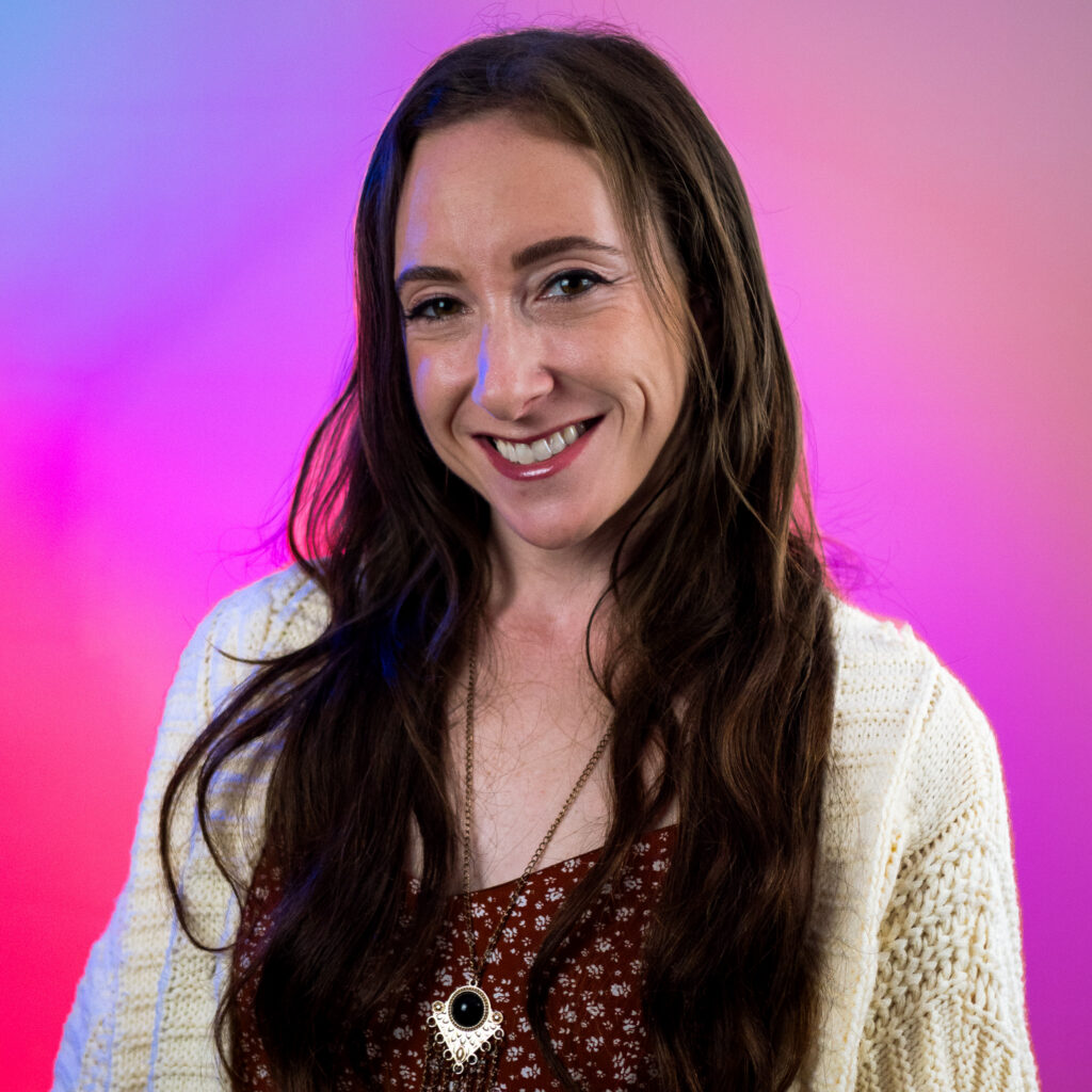 A woman with long brown hair smiles at the camera, wearing a cream knit cardigan over a red patterned top and an orsis pendant necklace. The background is a gradient of pink, purple, and blue.