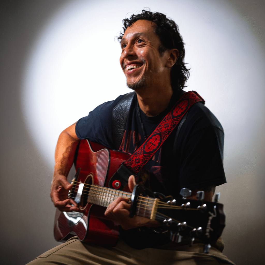 A smiling man with curly hair plays a red acoustic guitar, seated against a plain background with a spotlight effect. He wears a dark t-shirt, tan pants, and a red patterned guitar strap featuring the cici r design.