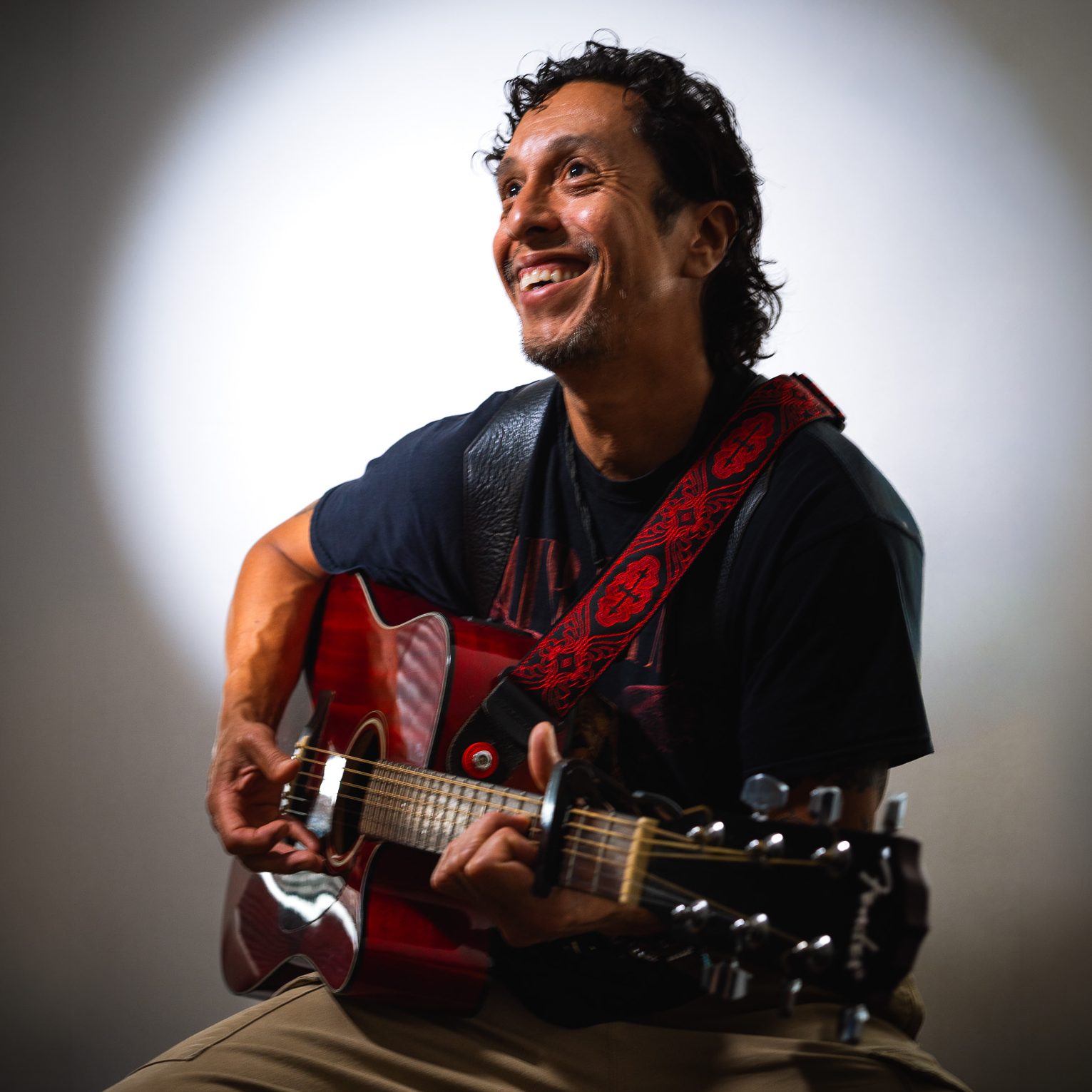 A smiling man with curly hair plays a red acoustic guitar, seated against a plain background with a spotlight effect. He wears a dark t-shirt, tan pants, and a red patterned guitar strap featuring the cici r design.