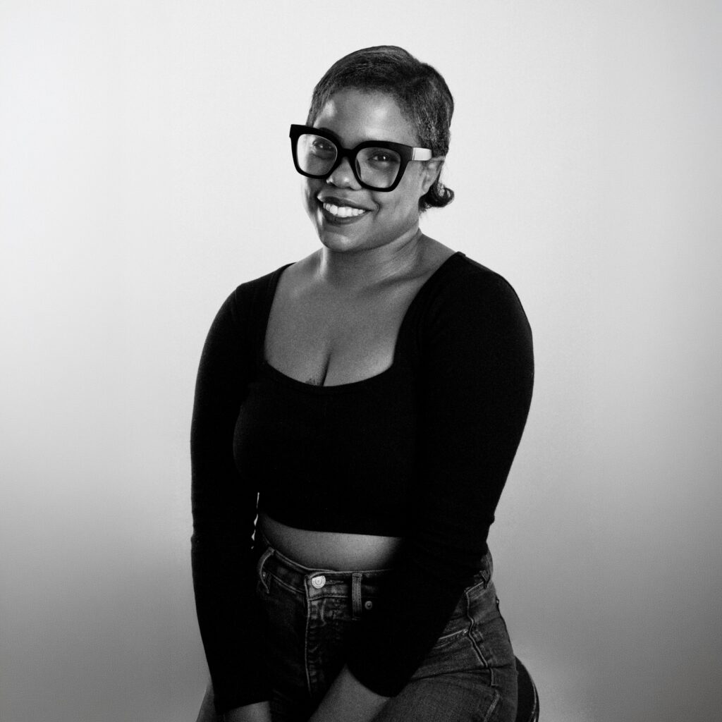 Mahalia C., a woman with short hair, wearing large glasses, a black long-sleeve crop top, and high-waisted jeans, smiles while sitting against a plain light background in a black-and-white photo.