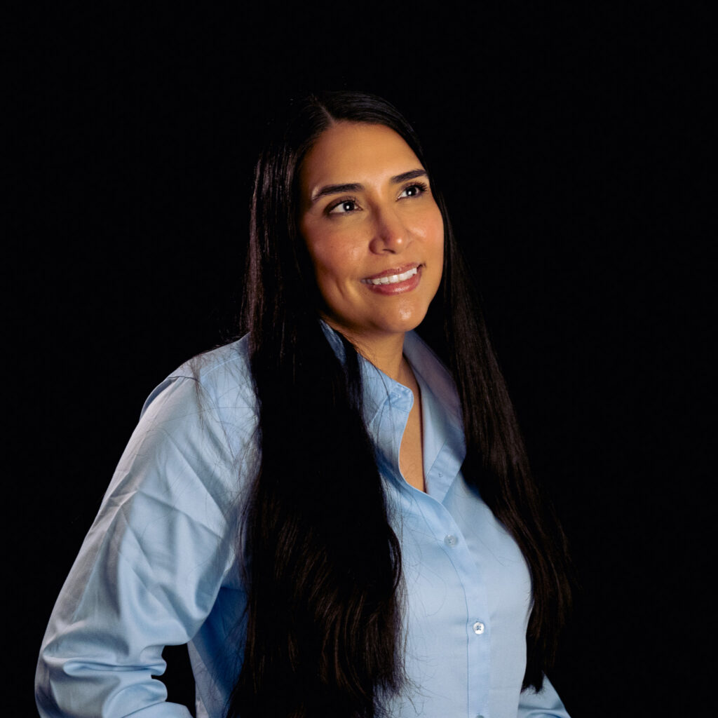 Carrianne M., a woman with long dark hair wearing a light blue button-up shirt, stands against a black background and smiles while looking slightly upward.