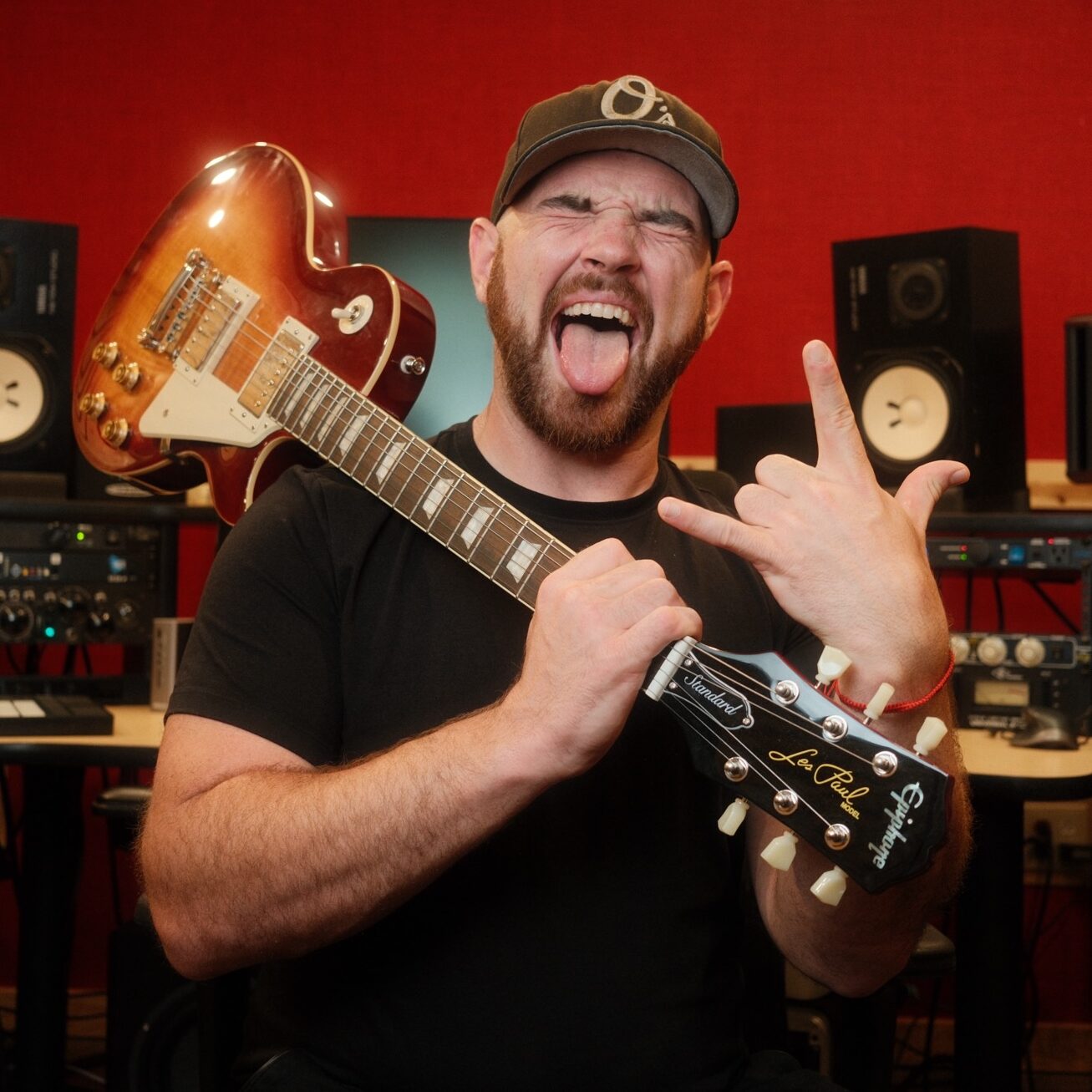 James P. stands in a recording studio, holding a Les Paul guitar over his shoulder, making a rock hand sign and sticking out his tongue. Dressed in black shirt and hat, he's surrounded by audio equipment and speakers.
