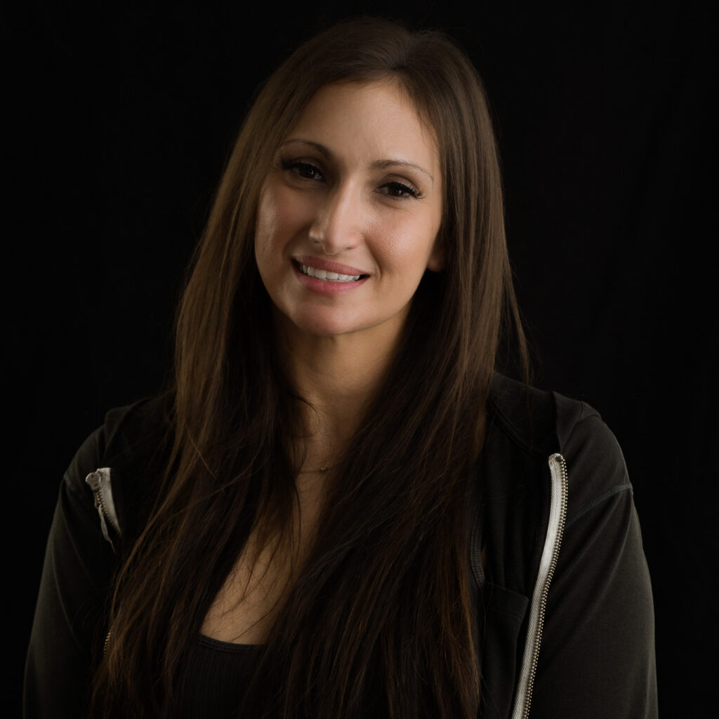 Lizzie T, a woman with long brown hair, wearing a black top and black zip-up hoodie, smiles softly against a dark background.