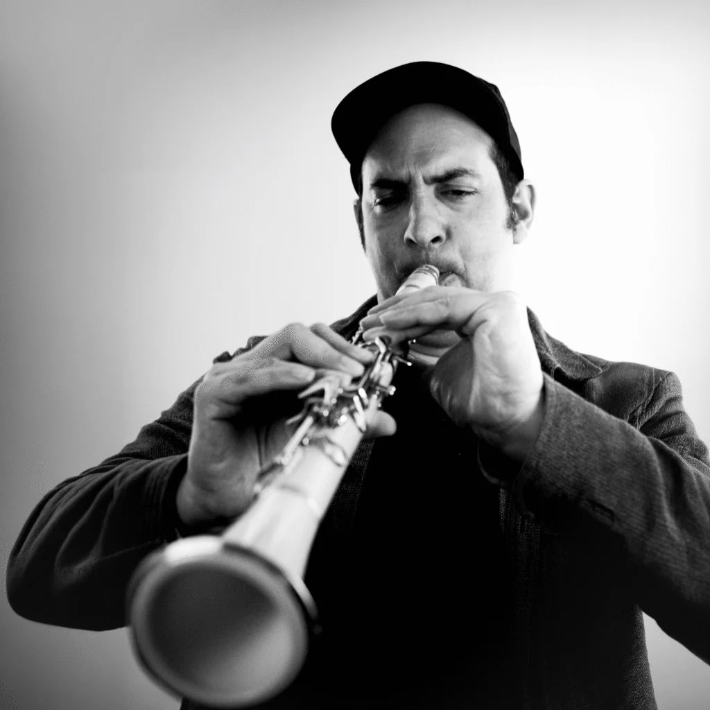 A man wearing a cap and a jacket plays a clarinet with intense focus against a plain, light background. The black-and-white image captures the kc musician’s dedication and passion for his craft.