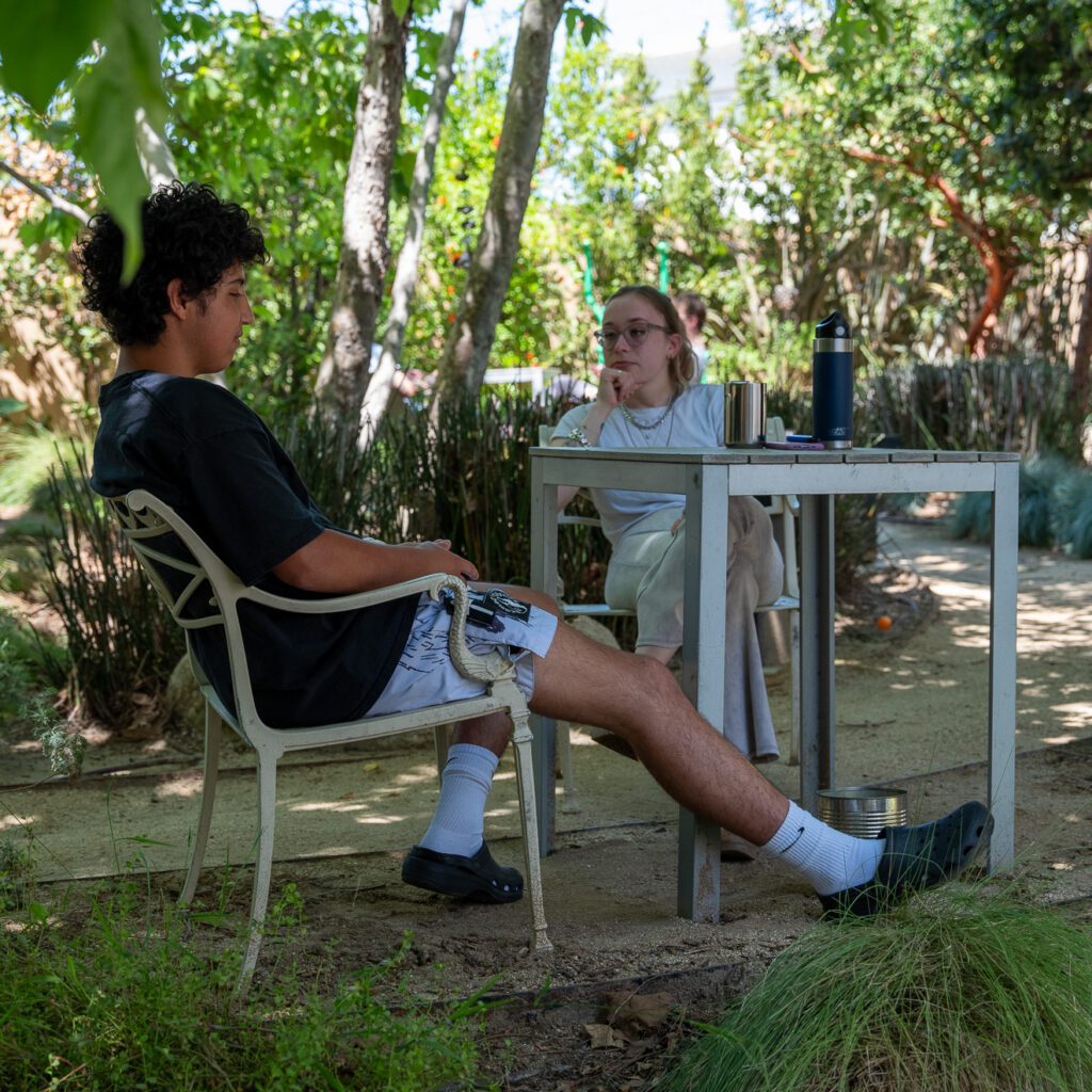 Two people sit at a small table outdoors in a lush, leafy garden. As sunlight filters through the trees above, their conversation hints at transformation—perhaps sharing stories of change found at a rehabilitation center near me.