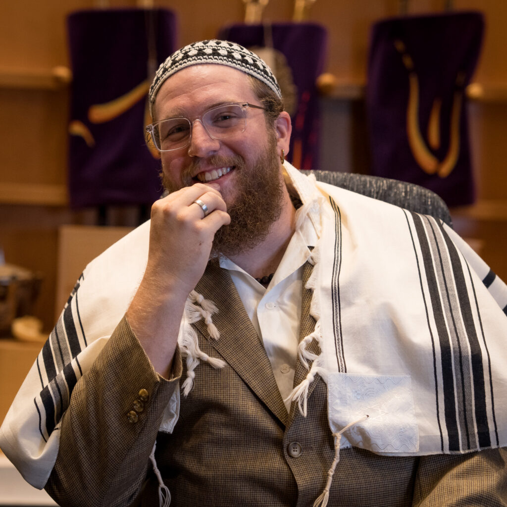 A smiling man, identified as David R, wears glasses, a patterned kippah, and a tallit draped over his suit while sitting indoors, with purple Torah scroll covers visible in the background.
