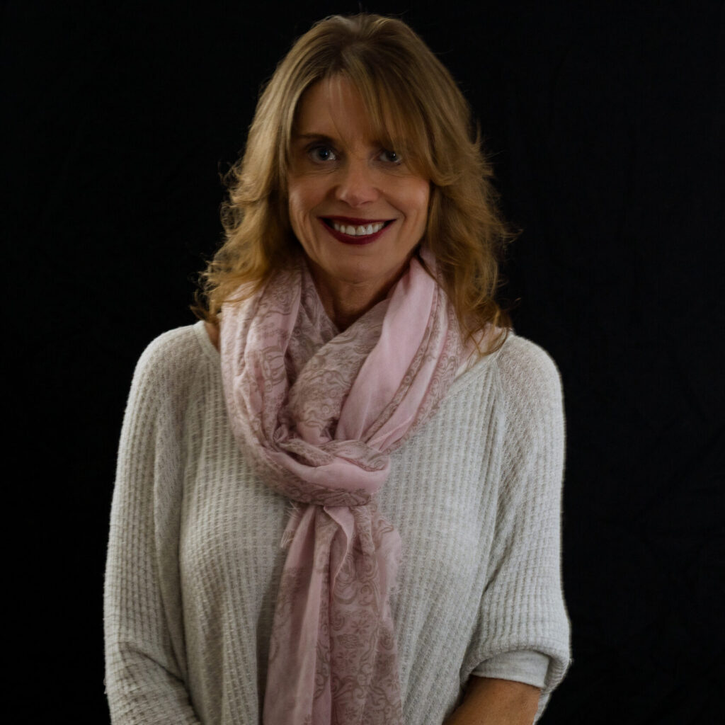 Lizzie, with shoulder-length light brown hair, wearing a white sweater and a pale pink scarf, smiles warmly while standing against a plain black background.