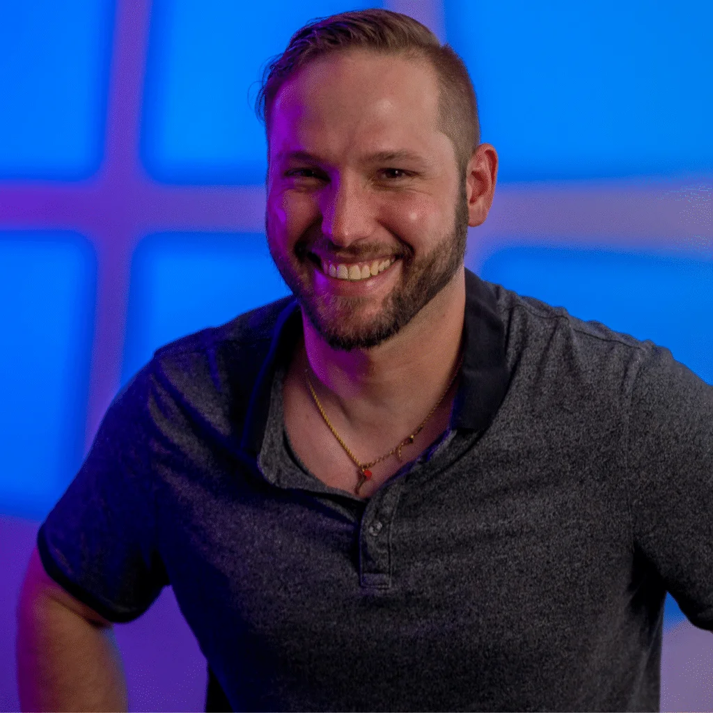 A smiling man with short hair and a trimmed beard, wearing a dark polo shirt and a necklace, poses in front of a blue and purple geometric background reminiscent of an Elaine N design.