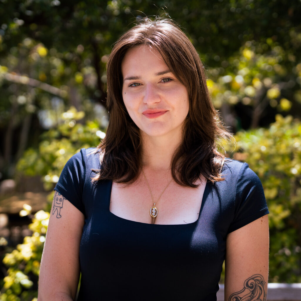 A woman with brown hair wearing a black top and gold necklace stands outdoors in sunlight, surrounded by greenery and trees. She has tattoos on her arms, a soft smile, and the portrait is shot in the distinctive style of Graeme N.