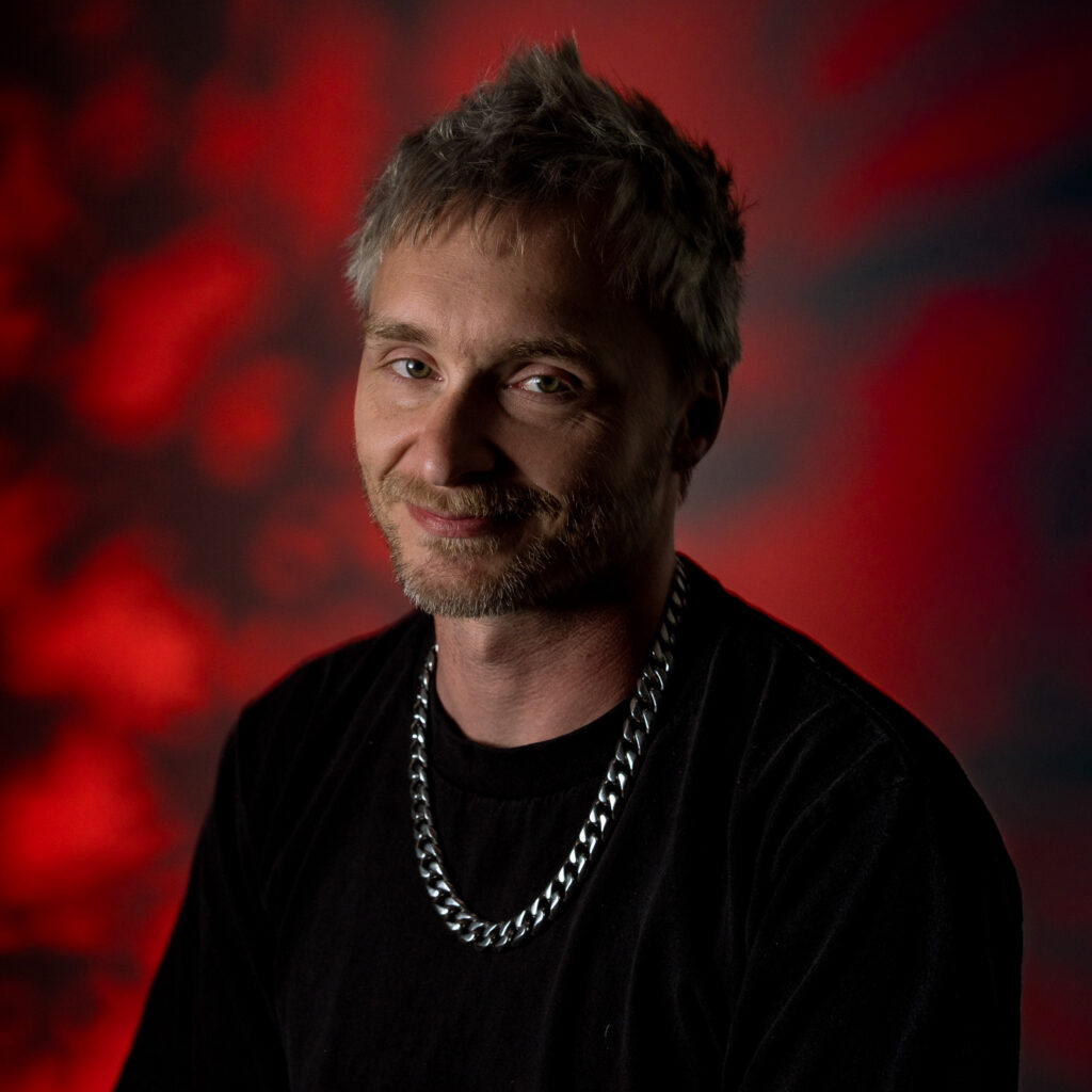 Owen H., a man with short, messy gray hair, a trimmed beard, and a silver chain necklace, smiles slightly. He wears a black shirt and stands against a red and black blurred background.