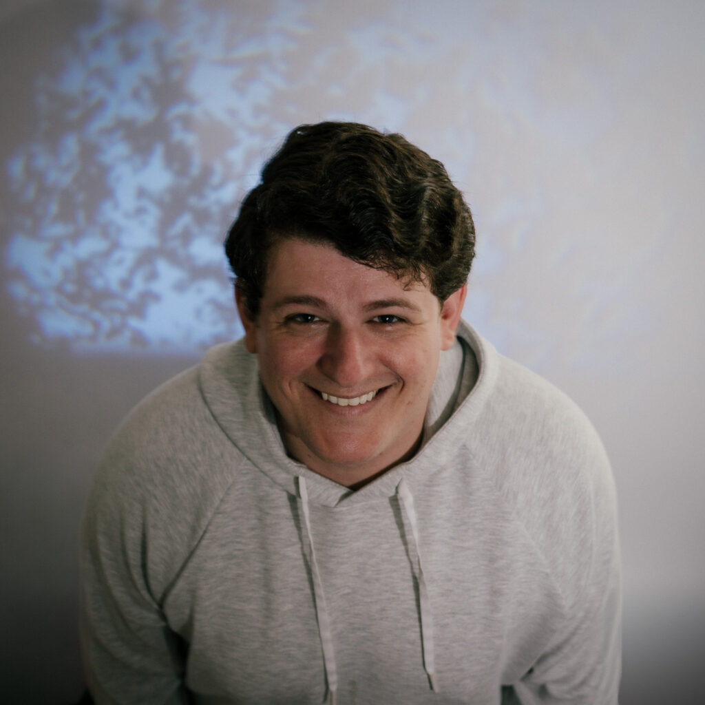 Mike C, a person with short, dark, wavy hair wearing a light gray hoodie, smiles at the camera with a blue and white abstract background behind them.