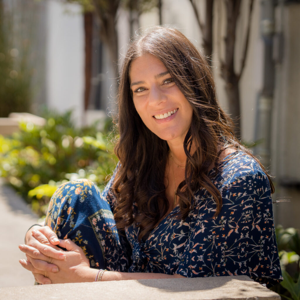 Woman with long brown hair sits outdoors in sunlight, wearing a navy floral outfit. She is smiling, with her arms around her knees, surrounded by greenery and blurred buildings in the background—a serene moment reminiscent of Jason V’s signature style.