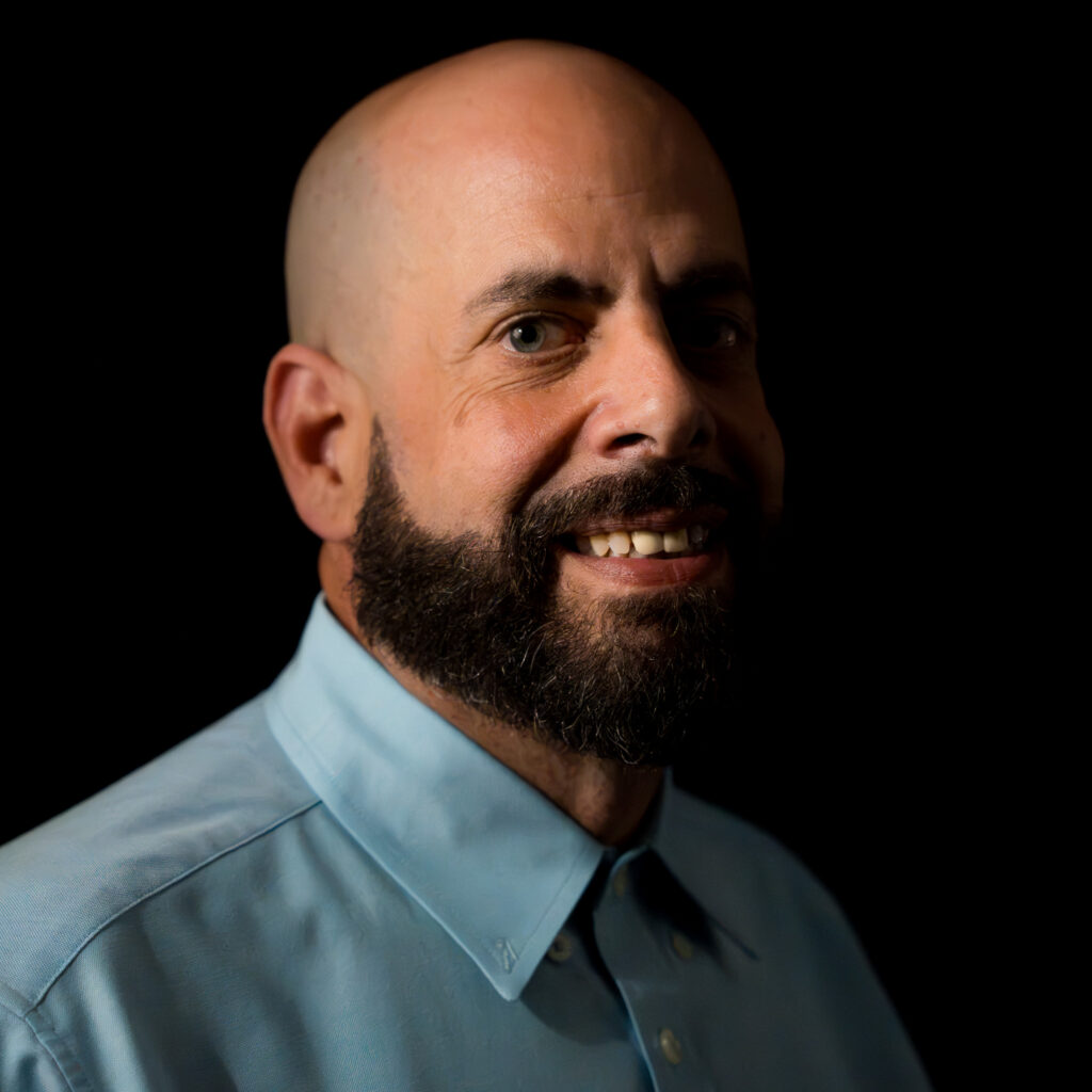 A man with a shaved head and full beard, identified as Josh G, wearing a light blue collared shirt, smiles against a black background.