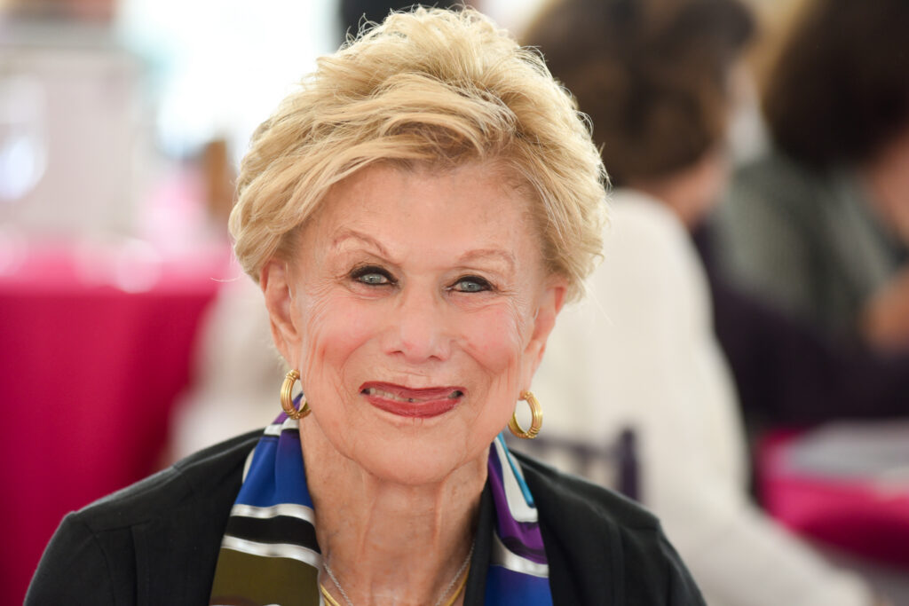 An elderly woman with short blonde hair smiles at the camera. She wears gold hoop earrings, a colorful scarf, and a black top. The background is softly blurred with pink tones and people sitting at tables. Annette Shapiro’s warmth radiates through the image, capturing the spirit of community fostered by the Legacy Fund.