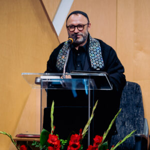 A man wearing glasses and a patterned scarf speaks at a clear podium with a microphone during events or programs, standing in front of a wooden wall and two chairs, with red flowers in front of him.