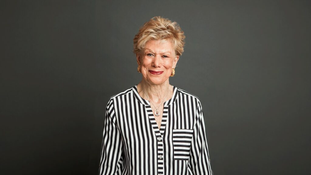 An older woman with short, light hair, wearing a black and white vertically striped shirt, stands smiling in front of a plain dark gray background.