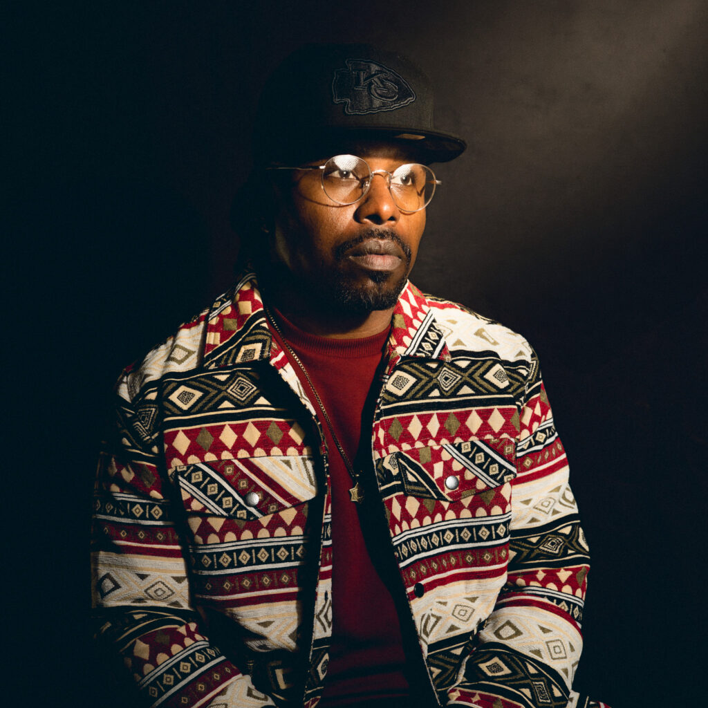 A man, reminiscent of Andre J, wearing glasses, a dark cap, and a patterned jacket with red, black, and white designs sits against a dark background, looking slightly away from the camera.