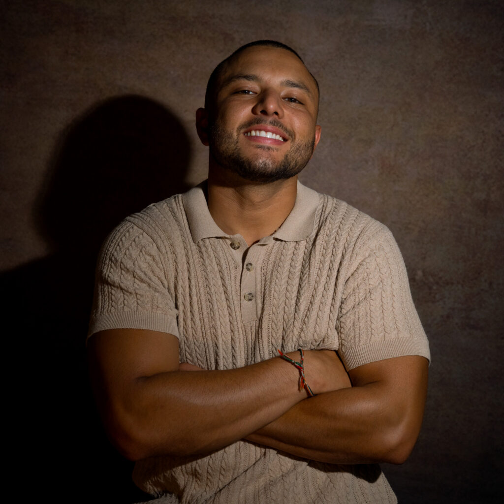 A smiling man with a trimmed beard and buzz cut poses with arms crossed, wearing a beige short-sleeve knit polo shirt, in front of a textured brown background—a confident portrait that perfectly reflects the approachable style of Jason C.
