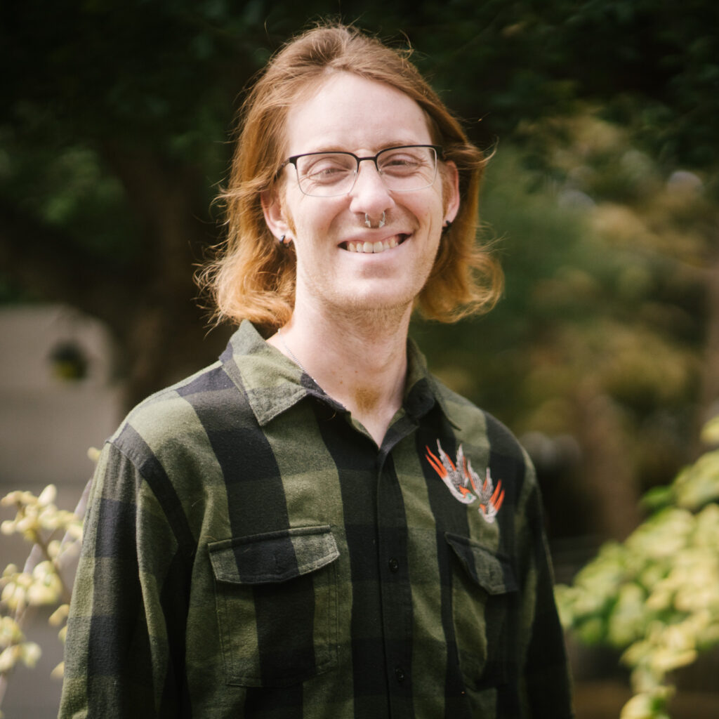 A person with glasses and shoulder-length light brown hair smiles outdoors, wearing a green and black plaid shirt with a bird design on the chest. Green foliage is visible in the blurred background, as rene p enjoys a peaceful moment in nature.