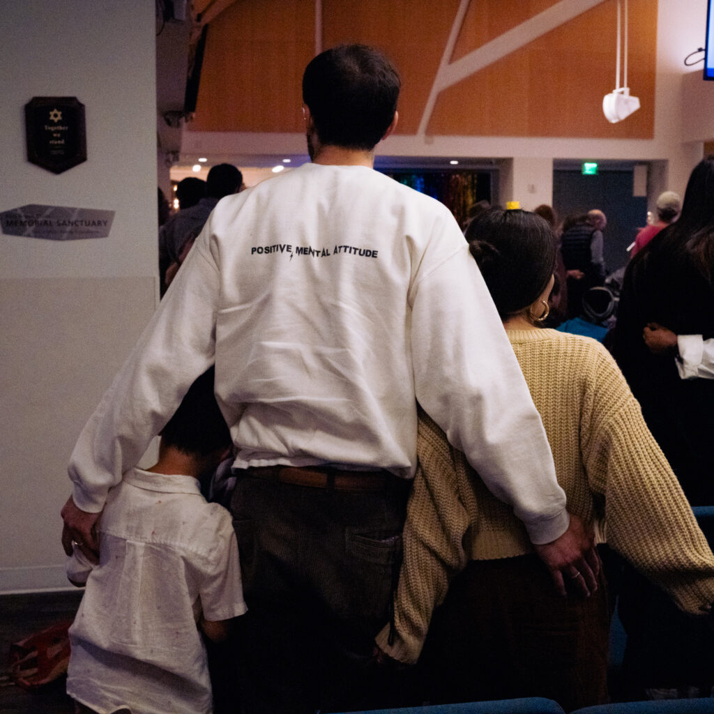 A man in a white sweatshirt reading “Positive, Mental Attitude” stands with his arms around two children in a crowded indoor setting, possibly a community event or gathering, embodying the spirit of mentorship and peer support.