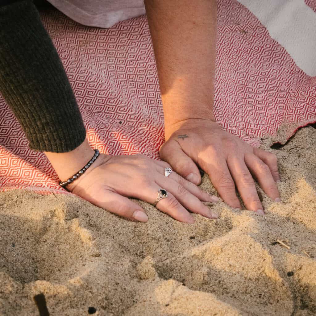 Two hands rest on a patterned red and white picnic blanket spread over beach sand, symbolizing the ongoing impact of family and addiction.